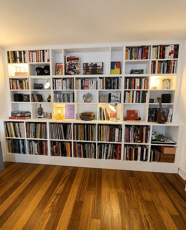 Bibliothèque blanche occupant tout un mur, casiers de tailles variées et bandeau bas pour vinyles.